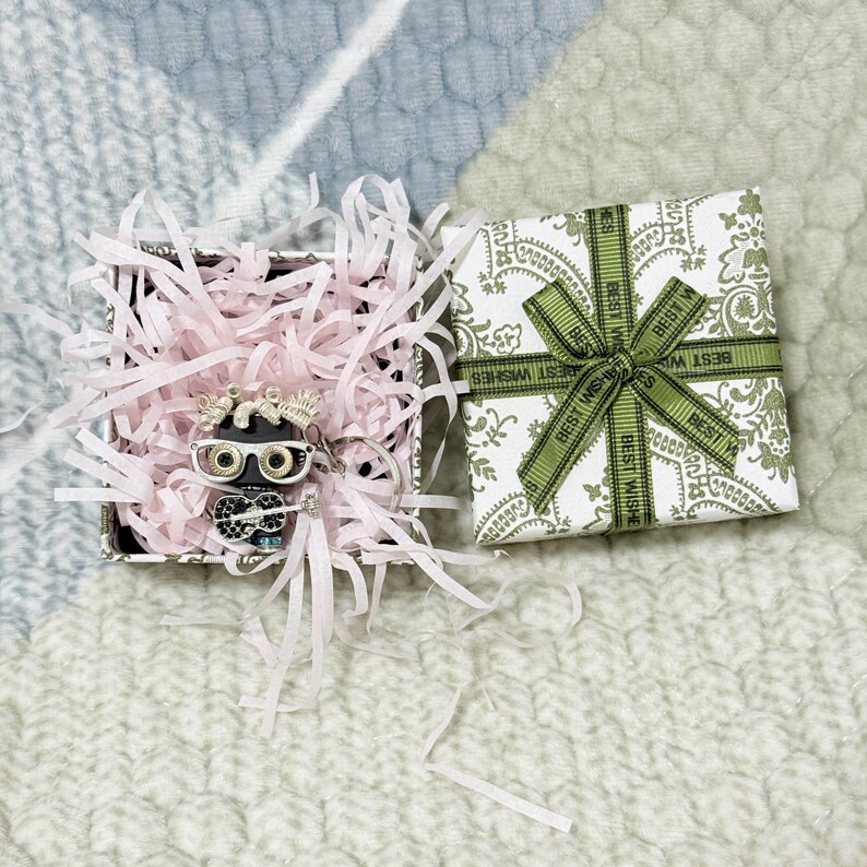 May include: A black and silver-toned owl-shaped keychain with clear and black rhinestones, wearing white-rimmed glasses and holding a guitar, nestled in a square gift box with pink paper shreds and a white lid featuring a green paisley print and ribbon.