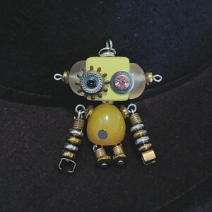 May include: A robot-shaped pendant crafted from mixed media components. The head is a yellow square with a gear-shaped eye, a pink rhinestone eye, and translucent side spheres. The body is a smooth amber-colored bead with a small gray dot.