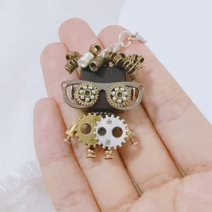 Puede incluir: Colgante de robot estilo steampunk con una cadena plateada. El robot presenta ojos en forma de engranaje, una cabeza negra y cabello rizado de metal. El cuerpo está hecho de engranajes dorados y plateados, con pequeños pies de engranaje.