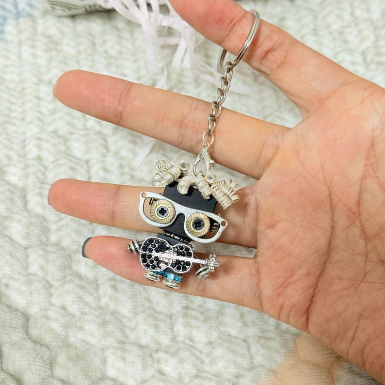 May include: A silver-toned metal keychain featuring a small robot figure. The robot has a black rectangular head, silver-rimmed glasses with gold-toned screw lenses, coiled silver wire hair, and a black body decorated with small black crystals.