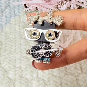 May include: A keychain featuring a small robot figure constructed from metal components. The robot has silver coiled wire hair, silver-rimmed glasses with screw-like lenses, and holds a black guitar encrusted with small dark crystals.