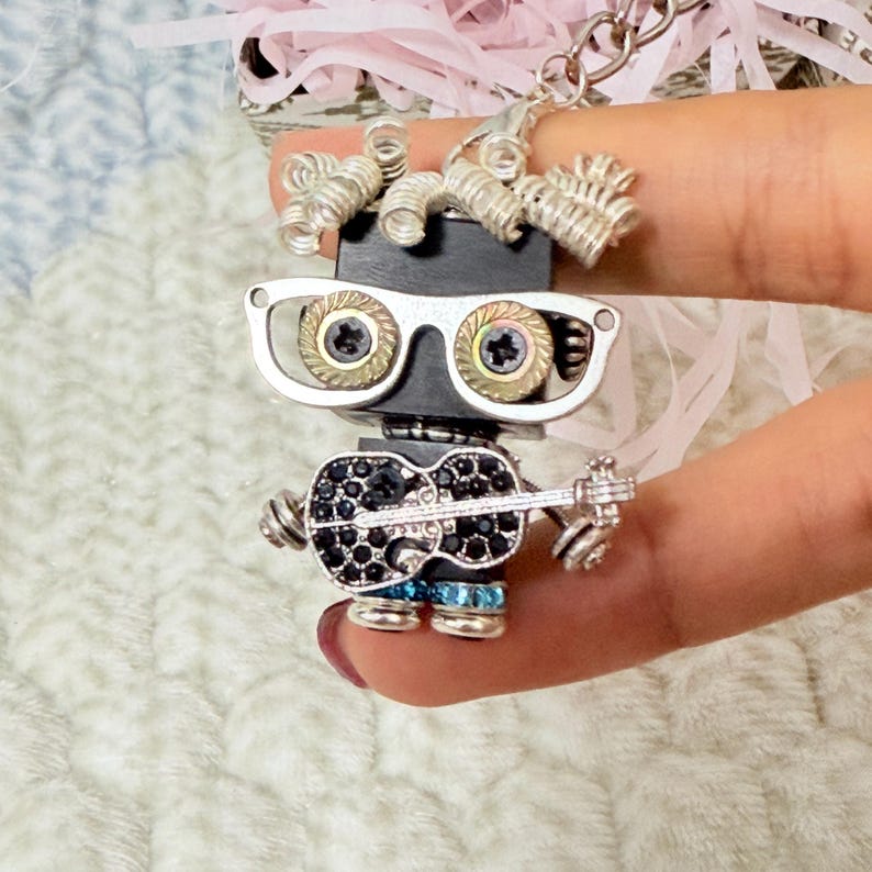 May include: A keychain featuring a small robot figure constructed from metal components. The robot has silver coiled wire hair, silver-rimmed glasses with screw-like lenses, and holds a black guitar encrusted with small dark crystals.