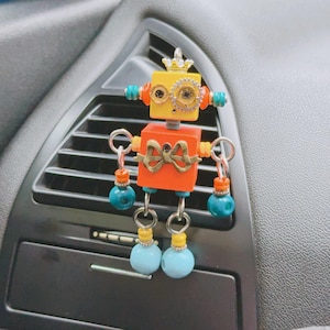 Könnte beinhalten: Ein farbenfroher Roboter-Auto-Lufterfrischer. Der Roboter hat einen gelben Kopf mit einer goldenen Krone und orangefarbenen Augen. Der Körper ist orange mit einer Schleife. Arme und Beine bestehen aus Metallringen und Perlen.
