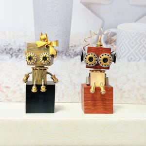 Op de afbeelding: Twee handgemaakte robotsculpturen. Eén robot heeft een gouden strik, zwarte ogen en een gouden lichaam. De andere robot heeft een rode kop, zilveren en gouden tandwielen voor ogen en een gouden lichaam. Beide robots zitten op houten blokken.