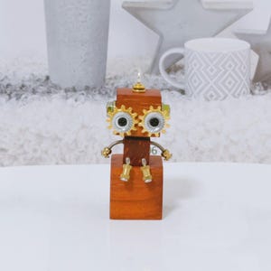 May include: A wooden robot ornament with cog-shaped eyes and metal detailing. The robot has a square body and head, with small metal legs and arms. The eyes are gold-coloured cogs with green and silver details. A unique decorative item.