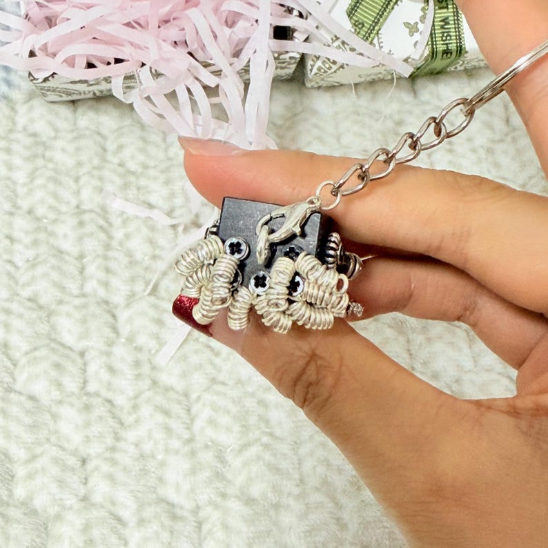 May include: A black cube-shaped keychain charm featuring multiple silver-toned metal springs attached to the surface with small screws. The charm is connected to a silver metal chain and a circular split ring, showcasing a mechanical design.