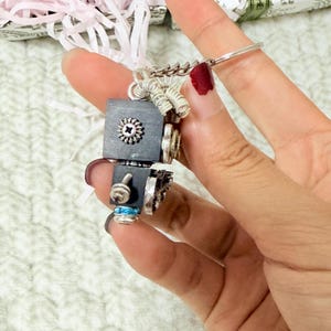 May include: A keychain featuring a small, black, cube-shaped component adorned with silver-toned metal hardware, including a screw-like detail, coiled wire accents, and a single bright blue bead, all attached to a standard silver metal keyring.