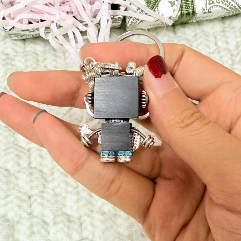 May include: A robot-shaped keychain featuring a square black head and torso, silver-toned metal arms and legs, and small blue rhinestones accenting the feet. The figure is attached to a circular silver metal split ring for keys.
