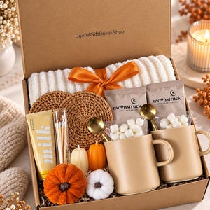 May include: A gift box from JoyfulGiftBoxesShop containing a white ribbed blanket with an orange ribbon, two tan mugs with gold spoons, two Moonstruck hot cocoa packets with marshmallows, two rattan coasters, a tube of milk lotion, and pumpkin candles.