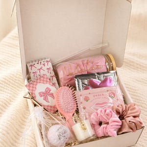 May include: A white gift box containing a glass tumbler with the name Valentina, a spiral notebook with pink bows, a pink hairbrush, a peach hydrating lip mask, two heart-shaped eye patches, a bath bomb, a small candle, and two fabric hair scrunchies.