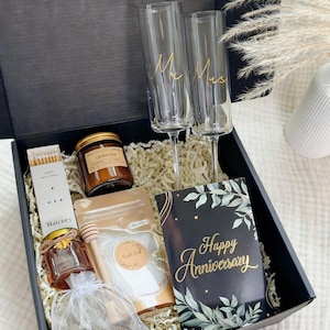 May include: A black gift box filled with items for a celebration. Includes two champagne flutes with "Mr Mrs" in gold script, a candle, matches, honey, bath salts, chocolates, and a card that says "Happy Anniversary".