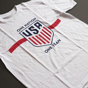 Pode incluir: Camiseta branca com um design de escudo com a inscrição "USA" em azul e vermelho, e as frases "ONE NATION" e "ONE TEAM". Uma faixa vermelha cruza a camiseta na diagonal.