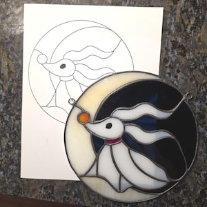 May include: A circular stained glass panel featuring a white ghost dog with long ears and a red collar, holding a small orange sphere. The design is set against a crescent moon background with cream and black glass segments and metal lead lines.