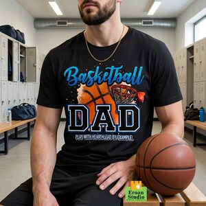 Puede incluir: Camiseta negra con un diseño de baloncesto y las palabras "Basketball DAD" en azul y blanco. La camiseta también incluye el texto "PLAY WITH HEART & LEAD BY EXAMPLE". Un balón de baloncesto marrón está en primer plano.