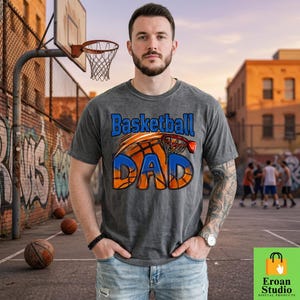 Puede incluir: Una camiseta gris con las palabras "Basketball DAD" en azul y naranja, con un diseño de baloncesto y aro. La camiseta la lleva una persona de pie en una cancha de baloncesto. Hay balones de baloncesto en el suelo.