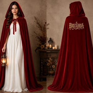 May include: A floor-length deep red velvet hooded cloak featuring a front ribbon tie closure. The back of the cloak displays the embroidered gold text "BRUXARIA" with a small crescent moon symbol above the lettering, presented in a full-length view.