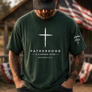 Puede incluir: Una camiseta de manga corta de color verde oscuro con un gráfico de cruz blanca sobre el texto FATHERHOOD IS KINGDOM WORK y PROVERBS 22:6. La manga izquierda muestra los nombres Sophia y Alex en letra blanca sobre una cruz pequeña.