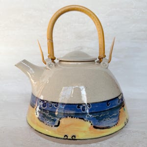 May include: A ceramic teapot with a light beige upper body and a curved bamboo handle. The lower section features a hand-painted design with a horizontal blue band depicting stylised waves above a yellow base with small black circular accents.