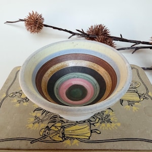 May include: A ceramic bowl featuring concentric circular bands of colour on the interior, including shades of yellow, light blue, brown, black, green, and pink. The exterior of the bowl is a solid off-white colour with a smooth, rounded rim.