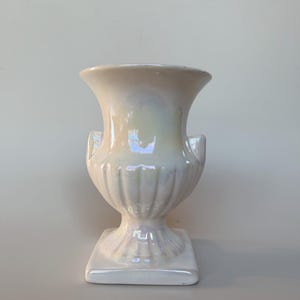 May include: A cream-coloured ceramic urn vase with an iridescent, pearlescent finish. The vase features a wide, flared rim, a bulbous body with vertical fluted detailing, two small side handles, and a square base. The surface reflects light.