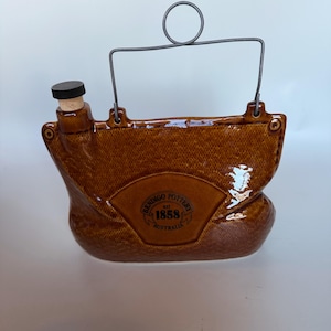 May include: A brown glazed ceramic canteen with a textured surface, a cork stopper with a black cap, and a grey wire handle. The front features a circular stamp that reads Bendigo Pottery Est 1858 Australia, centered within a fan-shaped design.
