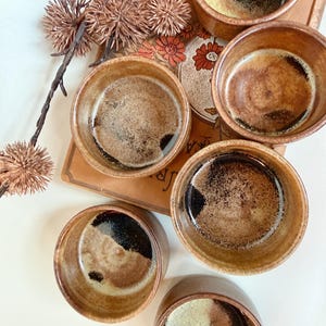 May include: A set of six round ceramic bowls with a speckled brown and tan glaze, featuring dark interior shading. The bowls are arranged around a rectangular book with a floral cover and a decorative branch with dried spiky seed pods.