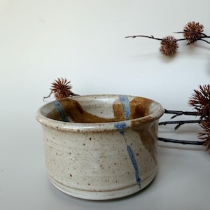 May include: A cylindrical ceramic bowl with a speckled cream-coloured glaze, featuring vertical streaks of brown and blue pigment that extend from the inner rim down the exterior wall, creating a textured appearance with a smooth, rounded top edge.
