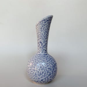 May include: A ceramic bud vase with a spherical base and a tall, slender neck. The surface features a textured, mottled pattern in shades of periwinkle blue and white, creating a speckled appearance across the entire exterior of the vessel.