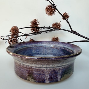 May include: A round ceramic bowl featuring a wide, flared rim and a deep basin. The glaze displays a mottled blend of deep purple, indigo, and earthy brown tones with a speckled texture throughout the exterior and interior surfaces.