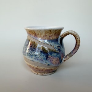 Peut inclure: Une tasse en céramique avec un corps arrondi et une anse incurvée. La surface présente un émail tacheté dans des tons de bleu, de beige et de brun, avec des bandes diagonales d'argile beige clair non émaillée et un intérieur blanc.