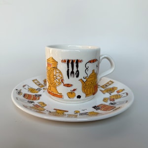 May include: A white ceramic espresso cup and matching saucer featuring a repeating pattern of orange and brown kitchen-themed illustrations, including a coffee grinder, a kettle, a rack of utensils, and various food items like apples.
