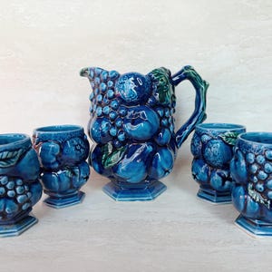 May include: A ceramic jug and four matching cups in a deep blue glaze. Each piece features a three-dimensional relief pattern of clusters of grapes and various fruits with green leaves. The jug has a curved handle and a pedestal base.