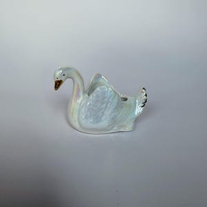 May include: A ceramic swan figurine featuring an iridescent, pearlescent finish that reflects light with subtle rainbow hues. The swan has a curved neck, a gold-toned beak, and detailed moulded wing feathers, with a small opening on its back.