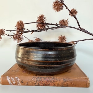May include: A round, dark brown ceramic bowl with a glossy, speckled finish and horizontal ridges along the exterior. The bowl rests on a tan, rectangular book titled Favourite Fairy Tales, which features a floral illustration on its cover.