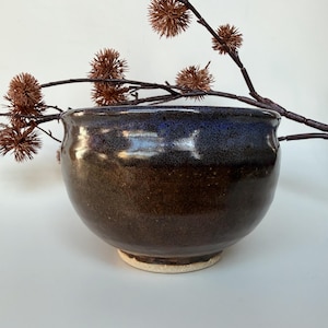 May include: A round ceramic bowl with a glossy finish, featuring a deep brown base that transitions into a dark blue rim. The bowl has a smooth, curved silhouette and sits on a light-coloured, unglazed circular base, accented by a brown twig.