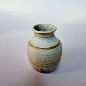May include: A small, rounded ceramic vase with a speckled off-white and beige finish. The vessel features a narrow neck, a flared rim, and a distinct horizontal brown band encircling the upper body, showcasing a textured, earthy appearance.