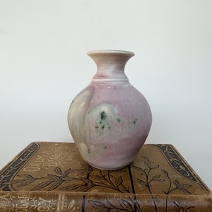 May include: A small, rounded ceramic vase with a flared rim, featuring a soft mauve and dusty pink glaze with mottled green and cream-coloured accents. The surface has a smooth, matte texture with subtle variations in colour throughout the body.