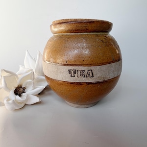 May include: A round, brown glazed ceramic canister featuring a wide, light-coloured horizontal band around the centre. The word TEA is embossed in dark, block-style lettering on the band. The canister has a thick rim and a matching ceramic lid.
