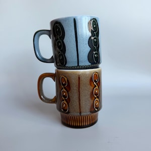 May include: Two stacked ceramic mugs featuring a speckled finish. The top mug is light blue with dark brown vertical accents and circular patterns. The bottom mug is brown with similar dark brown vertical accents and circular patterns, plus a ribbed base.