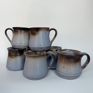 May include: A collection of six ceramic mugs featuring a two-tone glaze. Each mug has a light blue base with a speckled texture that transitions into a dark brown, smooth finish at the rim. The mugs have a rounded body and a curved handle.