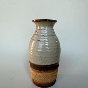 May include: A ceramic vase with a flared rim and a cylindrical body. The upper portion features a speckled off-white glaze, while the lower section transitions into a dark brown band followed by a textured, earthy tan base with horizontal ridges.