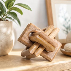 May include: A decorative sculpture crafted from light brown wood, featuring an interlocking geometric design composed of multiple square-shaped frames that are connected to form a complex, three-dimensional knot-like structure with visible grain.