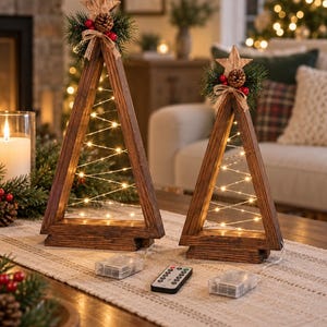 May include: Two triangular wooden frames shaped like Christmas trees, featuring warm white string lights wrapped around the interior. Each tree is topped with a wooden star, green pine needles, a small pinecone, and red berries, with battery packs and a remote.