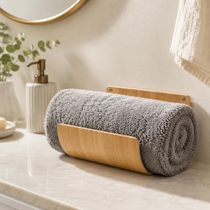 May include: A light-colored bamboo towel holder with a curved front panel and a flat back plate, mounted to hold a single rolled grey textured towel. The holder features visible metal screws at the top corners for secure wall attachment.