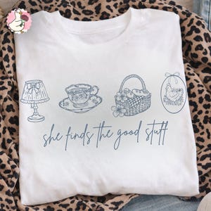 May include: White t-shirt with a blue line drawing of a lamp, teacup, basket, and an egg-shaped image of a chicken. The text "she finds the good stuff" is written in cursive below the drawings.