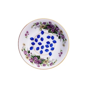 Puede incluir: Un plato de porcelana blanco con borde dorado y diseño floral, que contiene numerosos objetos pequeños, azules y de forma ovalada. El plato presenta patrones florales morados, amarillos y verdes.