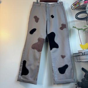 May include: Gray pants with patches of brown and black fabric sewn on. The pants are hanging on a hanger against a white wall. The patches are various shapes and sizes, adding a unique design element to the garment.