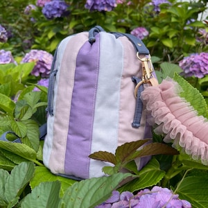 May include: A cylindrical fabric bag featuring vertical stripes in shades of purple, light lavender, and soft pink. The bag includes a dark blue strap attachment with a gold-toned metal swivel clasp and a decorative ruffled pink tulle accessory.