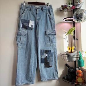 May include: Light blue denim cargo pants with patchwork details. The pants feature two cargo pockets and patches of various blue and black denim fabrics. The pants are hanging on a wooden hanger.