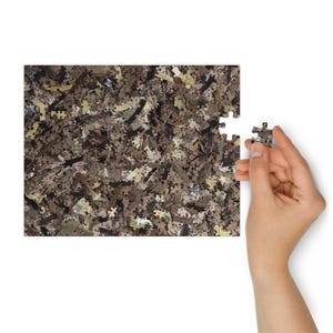 Peut inclure: Un puzzle partiellement assemblé avec un motif camouflage dans des tons de marron, beige et vert. Une main tient une seule pièce de puzzle, prête à être placée. Le puzzle est sur fond blanc.